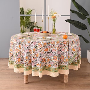 Peut inclure: Une nappe ronde avec un motif floral orange, rose et vert. La table est dressée avec des assiettes, des serviettes et des bougies. La nappe a une bordure rayée et est sur une table en bois.