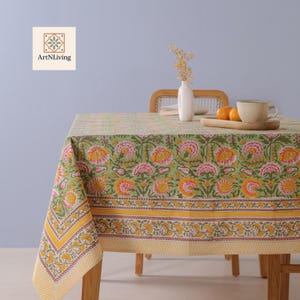May include: A green floral tablecloth with yellow and pink accents covers a wooden table. The tablecloth features a repeating pattern of large flowers and a yellow striped border. A wooden tray with oranges and a cup sits on the table.