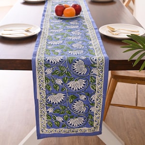 Op de afbeelding: Een blauwe tafelloper met bloemenpatroon met witte en groene bloemen en bladeren. De loper ligt op een donkere houten tafel, met borden, bestek en een fruitschaal.