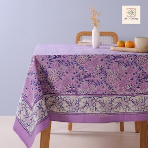 Może przedstawiać: Lawendowy obrus z kwiatowym wzorem w odcieniach fioletu i błękitu. Obrus ma dekoracyjne obramowanie z powtarzającym się kwiatowym wzorem. Na stole stoi biały wazon z suszonymi kwiatami.