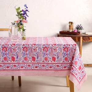 Könnte beinhalten: Ein rechteckiges weißes Tischtuch mit einem sich wiederholenden Blumenmuster in Rot- und Lilatönen. Das Tischtuch hat einen gestreiften Rand in Rot und Weiß. Eine Wasserkaraffe und Blumen stehen auf dem Tisch.
