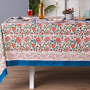 Puede incluir: Un mantel blanco con un estampado floral en rojo, naranja y verde, preparado para una comida. El mantel tiene un borde azul y está puesto con platos, servilletas y copas de vino. Una botella de vino y velas también están sobre la mesa.