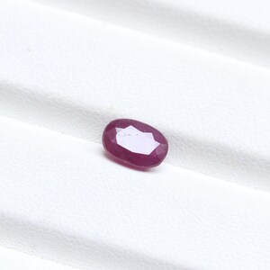 May include: A faceted, oval-shaped ruby gemstone. The gem is a deep, rich red color with a polished surface. The ruby is set against a white background, highlighting its color and clarity. The gemstone is a natural, untreated ruby.