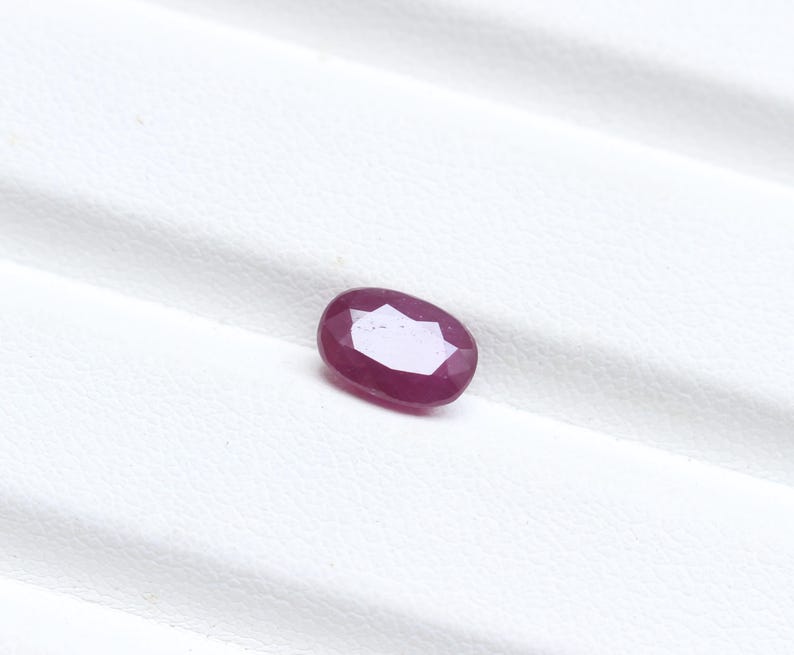 May include: A faceted, oval-shaped ruby gemstone. The gem is a deep, rich red color with a polished surface. The ruby is set against a white background, highlighting its color and clarity. The gemstone is a natural, untreated ruby.