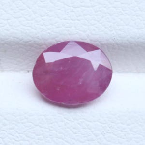 May include: An oval-shaped, faceted gemstone with a deep pinkish-red hue. The stone features a polished surface with multiple triangular and trapezoidal facets that reflect light, creating a clear, symmetrical pattern across the top of the gem.