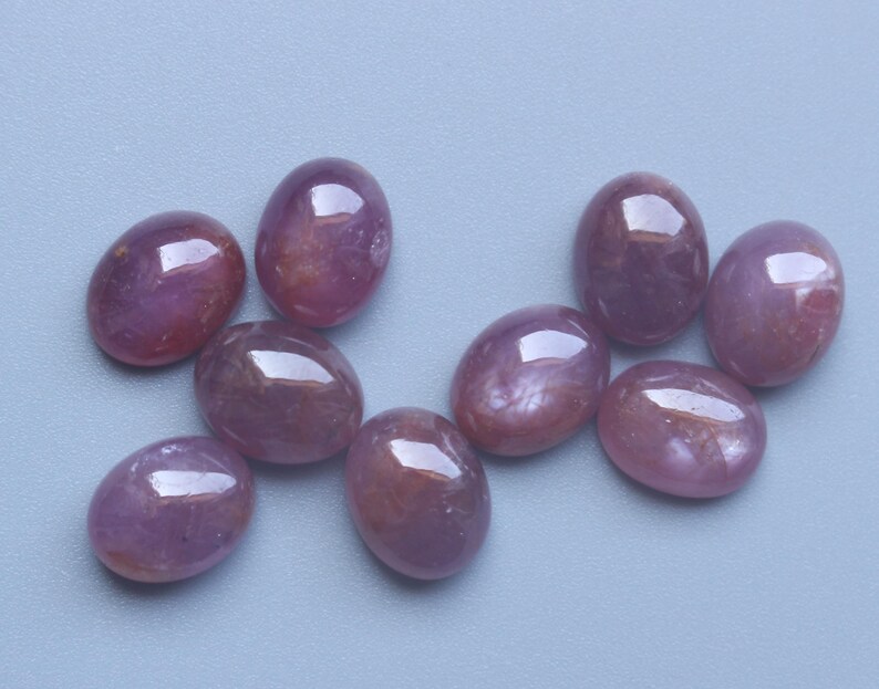 Pu&ograve; includere: Nove pietre preziose ovali e lucide in varie tonalit&agrave; di rosso-violaceo sono sparse su una superficie azzurra. Le gemme hanno una finitura liscia e lucida, che riflette la luce. Questi cabochon sono adatti per la creazione di gioielli.