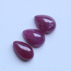 May include: Three polished, teardrop-shaped ruby gemstones. The gems are a deep, rich red colour with a glossy surface, set against a white background. The stones are arranged diagonally.