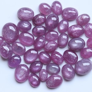 May include: A collection of oval-shaped, polished gemstones in varying shades of purple and pink. The gemstones have a smooth, glossy surface, reflecting light. The stones are scattered on a white surface, showcasing their natural beauty.