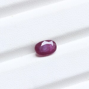 May include: A faceted, oval-shaped ruby gemstone. The ruby is a deep, rich red color with a slight purplish hue. The gemstone is set against a white background.