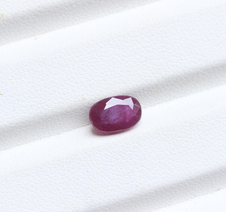 May include: A faceted, oval-shaped ruby gemstone. The ruby is a deep, rich red color with a slight purplish hue. The gemstone is set against a white background.