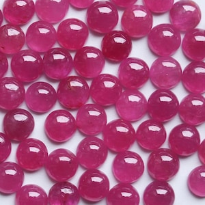 Könnte beinhalten: Eine Sammlung polierter, runder Rubin-Edelsteine. Die Edelsteine haben eine tiefe, lebendige, rosa-rote Farbe mit einer glatten, reflektierenden Oberfläche. Die Edelsteine sind über das Bild verstreut.