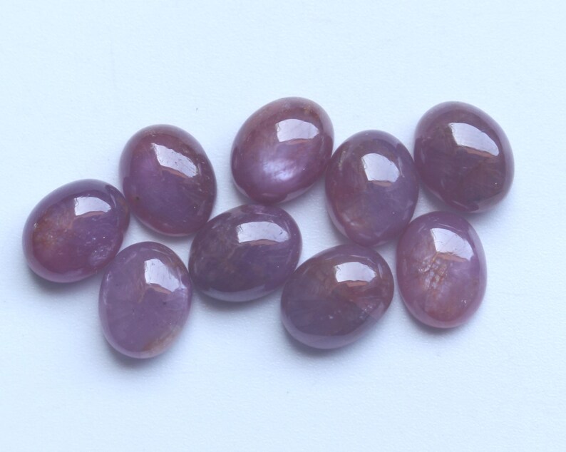 Pu&ograve; includere: Una collezione di nove pietre preziose ovali e lucide. Le pietre presentano una tonalit&agrave; viola intenso con sottili variazioni di colore e una superficie lucida e riflettente. Le pietre sono disposte su uno sfondo bianco.