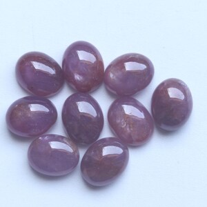 Pu&ograve; includere: Una collezione di nove pietre preziose ovali e lucide. Le pietre sono di un colore viola intenso con sottili variazioni e motivi interni. Ogni gemma ha una superficie liscia e riflettente.
