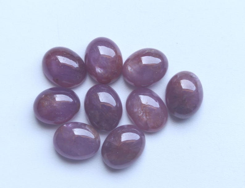 Pu&ograve; includere: Una collezione di nove pietre preziose ovali e lucide. Le pietre sono di un colore viola intenso con sottili variazioni e motivi interni. Ogni gemma ha una superficie liscia e riflettente.