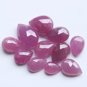 May include: A collection of faceted, teardrop-shaped gemstones in varying sizes. The gems are a deep pinkish-red color, with multiple facets catching the light. The arrangement is scattered, showcasing the gems' individual shapes and brilliance.