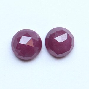 Könnte beinhalten: Zwei runde, facettierte Edelsteine in tiefroter Farbe. Die Edelsteine haben eine polierte Oberfläche mit einem geometrischen Schliff. Die Farbe ist ein sattes, dunkles Rot mit violetten Nuancen. Die Edelsteine sind vor einem weißen Hintergrund platziert.