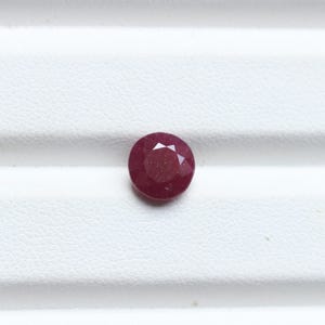 K&ouml;nnte beinhalten: Ein runder, facettierter Rubin-Edelstein. Der Edelstein hat eine tiefrote Farbe und liegt auf einer wei&szlig;en Oberfl&auml;che. Die Facetten erzeugen ein Sternmuster, das Licht reflektiert. Dieser Edelstein eignet sich f&uuml;r die Schmuckherstellung.