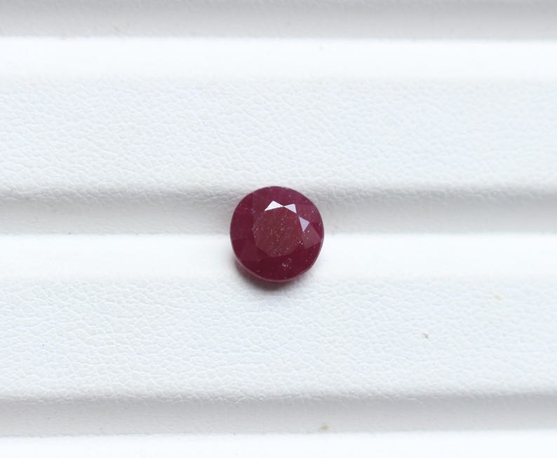 K&ouml;nnte beinhalten: Ein runder, facettierter Rubin-Edelstein. Der Edelstein hat eine tiefrote Farbe und liegt auf einer wei&szlig;en Oberfl&auml;che. Die Facetten erzeugen ein Sternmuster, das Licht reflektiert. Dieser Edelstein eignet sich f&uuml;r die Schmuckherstellung.