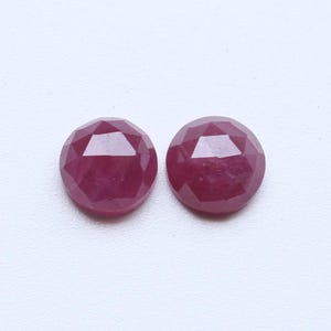 May include: Two round, faceted ruby gemstones. The rubies are a deep, rich red colour and have a polished, reflective surface. The gemstones are set against a white background.