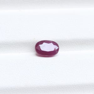 May include: A faceted, oval-shaped ruby gemstone. The gem is a deep, rich red color with multiple facets that reflect light. The ruby is set against a white background, highlighting its color and clarity. This gemstone is suitable for jewelry.
