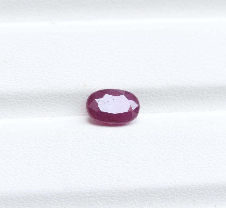 May include: A faceted, oval-shaped ruby gemstone. The gem is a deep, rich red color with multiple facets that reflect light. The ruby is set against a white background, highlighting its color and clarity. This gemstone is suitable for jewelry.