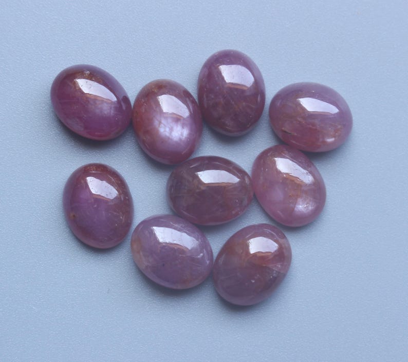 Pu&ograve; includere: Nove pietre preziose ovali e lucide in varie tonalit&agrave; di rosa e rosso sono disposte su una superficie azzurra. Le pietre preziose hanno una finitura liscia e lucida, che riflette la luce. Le pietre sembrano essere cabochon.