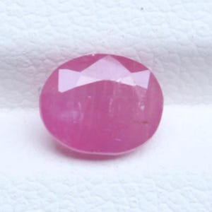 Könnte beinhalten: Ein leuchtender, ovaler rosa Edelstein mit einer facettierten Oberfläche, die durch mehrere geometrische Schliffe das Licht reflektiert. Der Stein zeigt einen gleichmäßigen, gesättigten magentaroten Farbton über seinen gesamten polierten, durchscheinenden Körper.