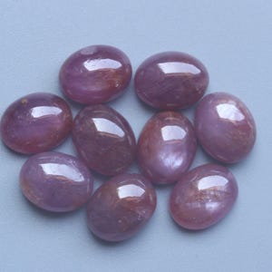 Pu&ograve; includere: Una collezione di nove gemme ovali e lucide. Le pietre sono di un colore rosso-violaceo intenso con una superficie lucida e riflettente. Sono disposte in un motivo circolare su uno sfondo azzurro chiaro.