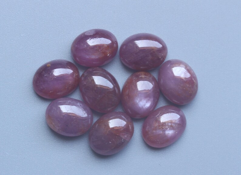 Pu&ograve; includere: Una collezione di nove gemme ovali e lucide. Le pietre sono di un colore rosso-violaceo intenso con una superficie lucida e riflettente. Sono disposte in un motivo circolare su uno sfondo azzurro chiaro.