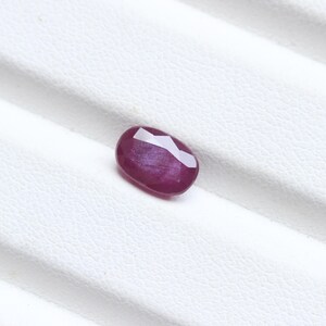 May include: A faceted, oval-shaped ruby gemstone. The gem is a deep, rich red color with a polished surface. The ruby is set against a white background.