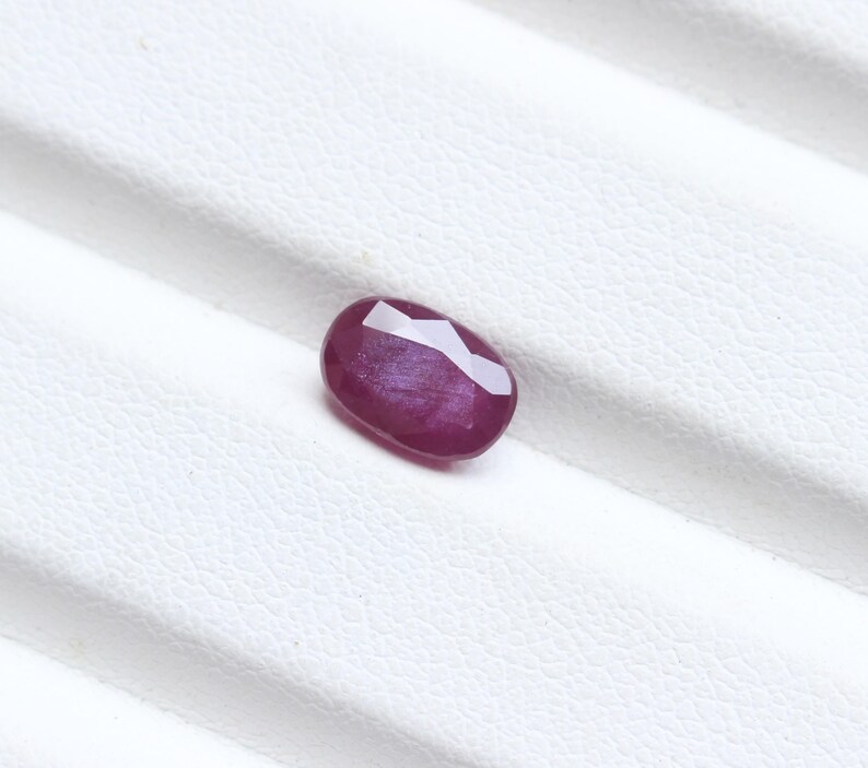 May include: A faceted, oval-shaped ruby gemstone. The gem is a deep, rich red color with a polished surface. The ruby is set against a white background.