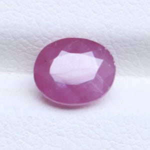 May include: A loose oval-shaped gemstone with a faceted surface and a vibrant pink color. The stone features a polished finish with visible internal inclusions and a clear, reflective center facet that catches the light.