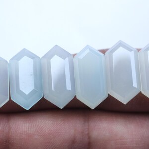 May include: Six pale blue, translucent, hexagonal-shaped gemstones with faceted edges. Each stone features a smooth, polished surface and a geometric cut, arranged in a horizontal row to display their uniform shape and light-reflecting properties.