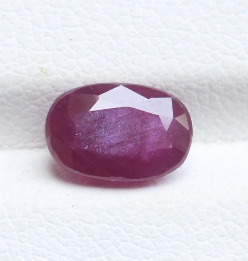 May include: A faceted, oval-shaped ruby gemstone. The gem is a deep, rich red color with a polished surface. The facets create a play of light and shadow, enhancing its brilliance. The ruby is set against a white background.