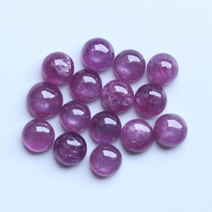 Pode incluir: Uma coleção de pedras preciosas redondas e polidas em tons de rosa e roxo. As pedras lapidadas em cabochão têm uma superfície lisa e brilhante. Estão espalhadas sobre um fundo branco. Adequadas para a criação de joias.
