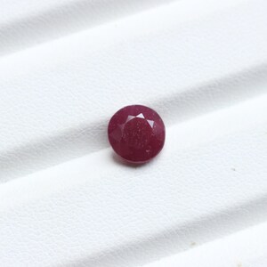 K&ouml;nnte beinhalten: Ein facettierter, runder Rubin-Edelstein. Der Edelstein hat eine tiefe, satte rote Farbe und scheint poliert zu sein. Der Rubin ist vor einem wei&szlig;en Hintergrund platziert, der seine Farbe und Form hervorhebt. Dieser Edelstein eignet sich f&uuml;r die Schmuckherstellung.