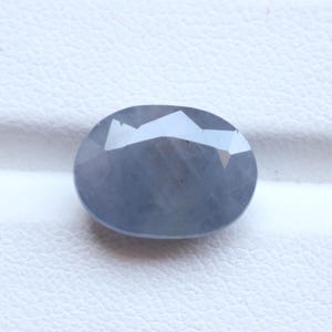 May include: A faceted, oval-shaped, blue-gray gemstone. The gem has a polished surface with multiple facets that reflect light. The stone is set against a white background, highlighting its color and shape. This gemstone could be used in jewelry.