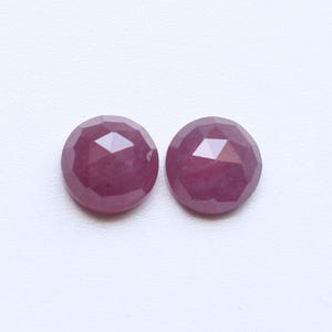 May include: Two round, faceted gemstones with a deep reddish-pink hue. The gems are set against a white background, showcasing their polished surfaces and geometric cuts. These gemstones are likely rubies, perfect for jewelry making.