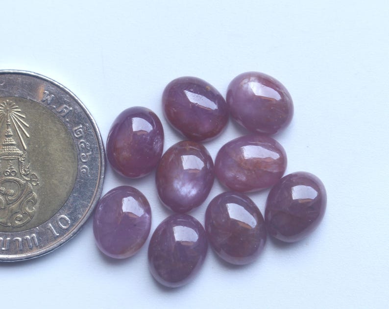 Pu&ograve; includere: Una collezione di nove gemme ovali, lucide, rosso-violacee. Le gemme sono disposte su una superficie bianca accanto a una moneta. Le gemme hanno una superficie liscia e riflettente e variano leggermente per colore e tonalit&agrave;.
