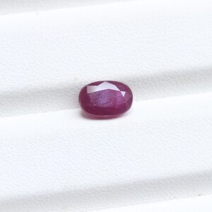 May include: A faceted, oval-shaped ruby gemstone. The gem is a deep red color with a slight purplish hue. The cut of the stone creates multiple reflective surfaces, enhancing its brilliance. The ruby is set against a white background.