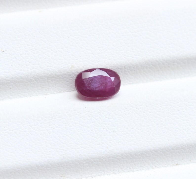 May include: A faceted, oval-shaped ruby gemstone. The gem is a deep red color with a slight purplish hue. The cut of the stone creates multiple reflective surfaces, enhancing its brilliance. The ruby is set against a white background.