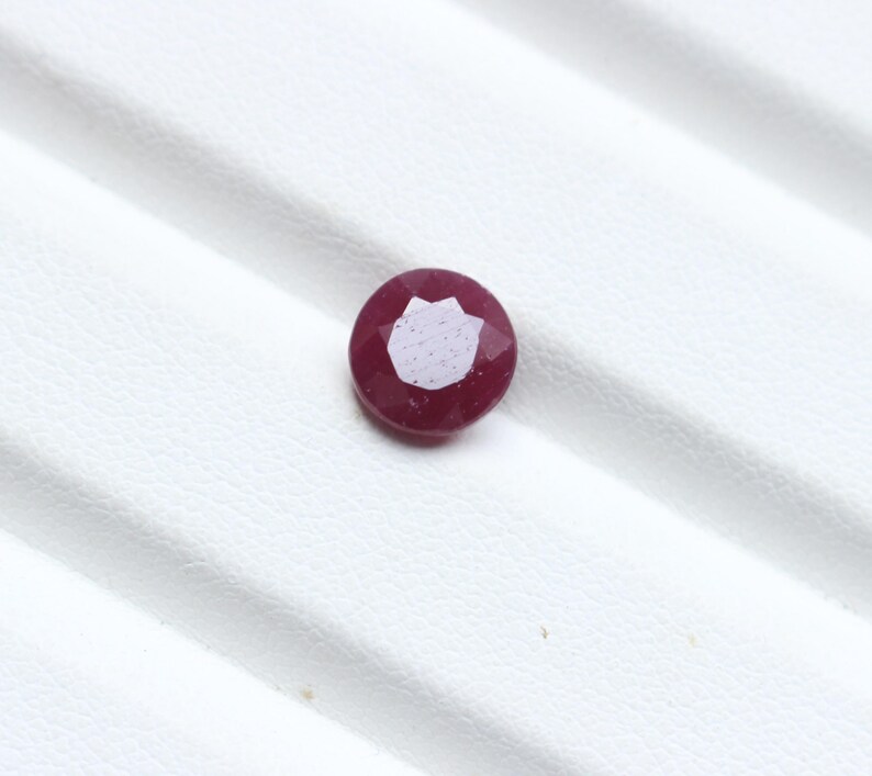 K&ouml;nnte beinhalten: Ein facettierter, runder Rubin-Edelstein. Der Edelstein hat eine tiefrote Farbe mit einem helleren, reflektierenden Zentrum. Der Stein liegt vor einem wei&szlig;en Hintergrund, der seine Farbe und seinen Schliff hervorhebt.