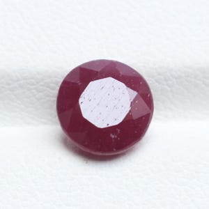 May include: A round, faceted ruby gemstone. The deep red gem has a bright, reflective surface. The facets create a geometric pattern. The stone is set against a white background.