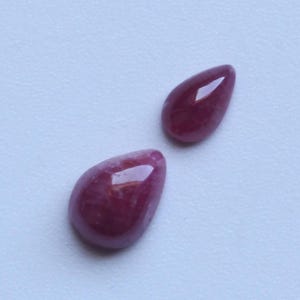 May include: Two polished, teardrop-shaped gemstones in a deep, rich red hue. The stones have a smooth, reflective surface, catching the light and displaying subtle variations in color. The gemstones are set against a plain, light background.