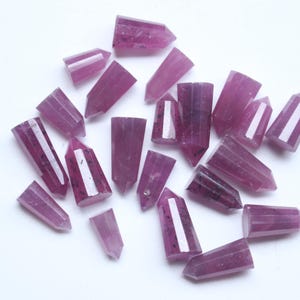 May include: A collection of small, polished, point-shaped crystals in varying shades of deep magenta and purple. Each piece features a faceted, hexagonal column structure with a pointed tip, displaying natural internal inclusions and smooth surfaces.