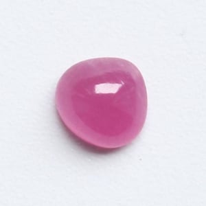 Puede incluir: Una gema pulida, de forma ovalada, en un vibrante tono magenta. La gema tiene una superficie brillante, que refleja la luz, y está sobre un fondo blanco liso. Podría ser un rubí tallado en cabujón u otra piedra preciosa.