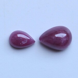 May include: Two polished, teardrop-shaped gemstones in a deep magenta hue. The stones have a smooth, reflective surface, with subtle variations in color and internal patterns. The gems are set against a light background.