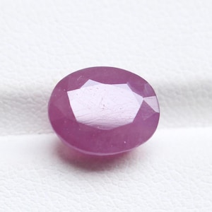 Pode incluir: Uma gema de rubi facetada, em forma oval. A gema é de cor vermelho-rosada profunda, com múltiplas facetas que capturam a luz. A pedra preciosa está sobre um fundo branco.