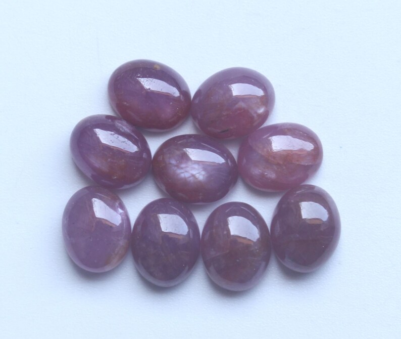 Pu&ograve; includere: Una collezione di otto pietre preziose ovali e lucide. Le pietre sono di un colore rosso-violaceo intenso con una superficie lucida e riflettente. Le pietre preziose sono disposte su uno sfondo bianco, mostrando le loro forme lisce e arrotondate. Adatto per gioielli.