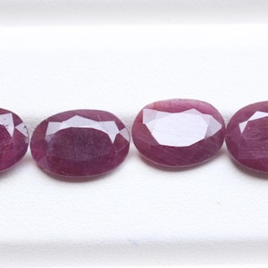 May include: Four oval-shaped, faceted rubies with a deep magenta-red hue. Each gemstone features a polished surface with multiple triangular facets that reflect light, arranged in a horizontal row against a plain, light-colored background.